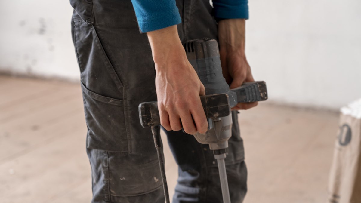 Handwerker in Arbeitshose hält einen Bohrhammer, im Hintergrund ein leerer Raum im Ausbau
